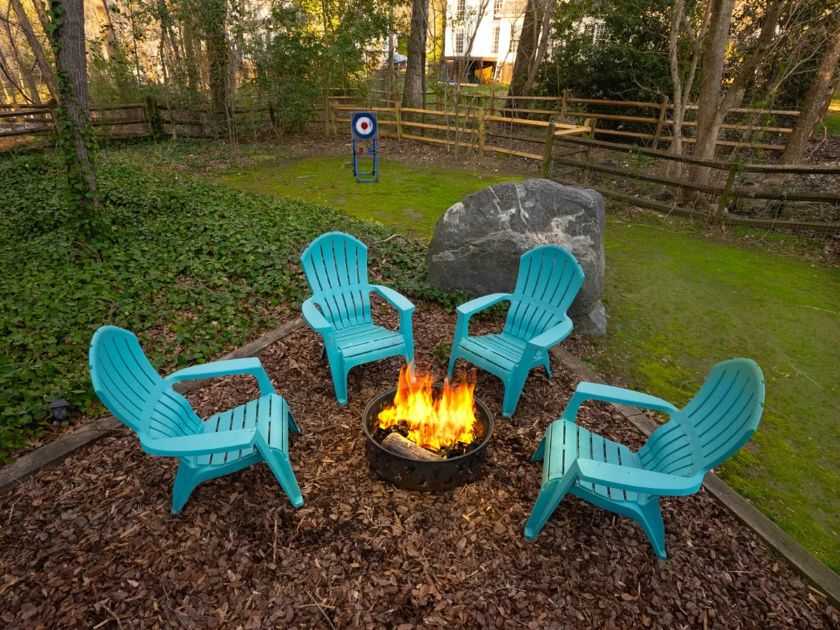 An outdoor fire pit area with turquoise chairs arranged in a circle, a large rock, and an archery target in the background. The setting is designed for communal gatherings, evening barbecues, and outdoor games.