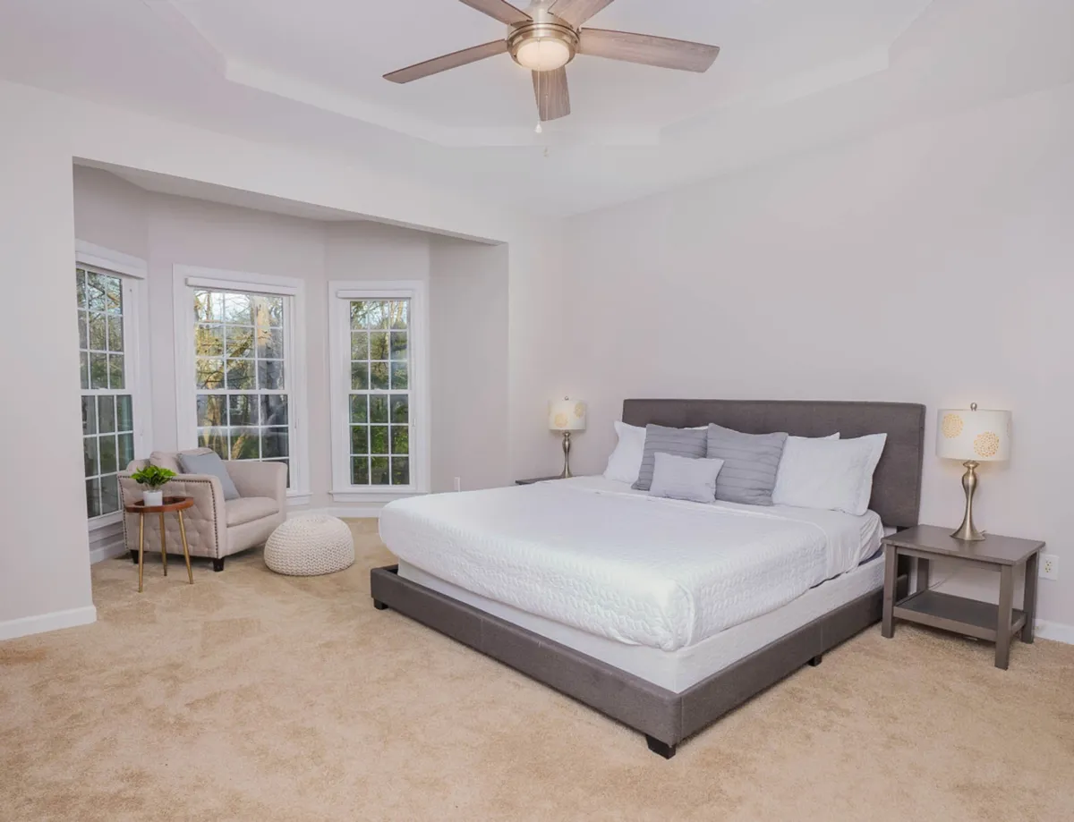 A spacious bedroom featuring a large bed with a gray headboard, an inviting armchair in the corner, ample natural light streaming through generous windows, and a small side table with a lamp. The combination of soft textiles and serene colors creates a restful and welcoming ambiance.