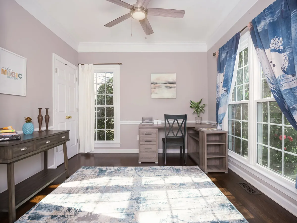 A flexible office space with a desk positioned by a large window, accented by artwork, a cozy rug, and ample natural light. The scene is calm and productive, ideal for working, reading, or transforming into a supplementary sleeping area as needed.