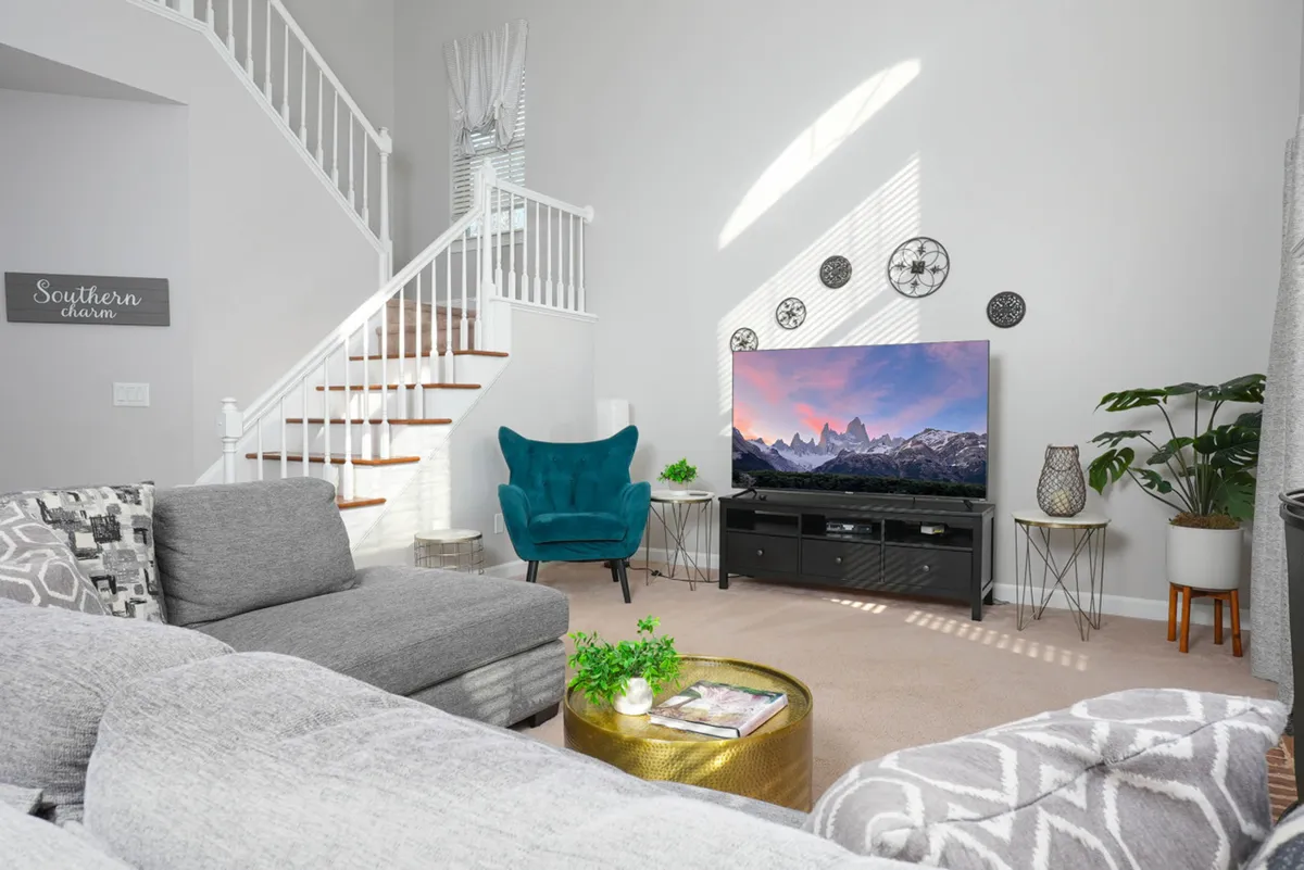 A cozy, open-concept living room featuring a large gray sectional sofa, a teal accent chair, a big-screen television, and a round coffee table with greenery. The space is designed for relaxation and socializing, with a visible staircase adding to the sense of openness.