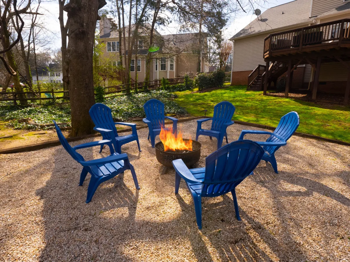 This image showcases a wood-burning fire pit surrounded by blue chairs, set in lush greenery, inviting guests to gather for storytelling, s'mores, and stargazing. The arrangement and lighting convey a warm, communal atmosphere perfect for evening gatherings.