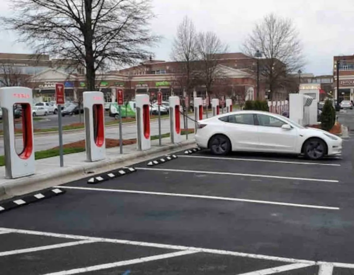 Tesla charging stations in an outdoor shopping area, with a white Tesla parked and visible retail spaces in the background. The scene is modern and accessible, indicating connectivity to local amenities and ease of travel.