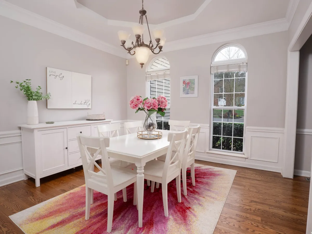 The image showcases a bright, spacious dining area with an elegant chandelier illuminating a large table set with a beautiful floral arrangement. The scene is inviting, with a cozy rug underfoot and plenty of room for guests, evoking the warmth of family dinners and festive gatherings.